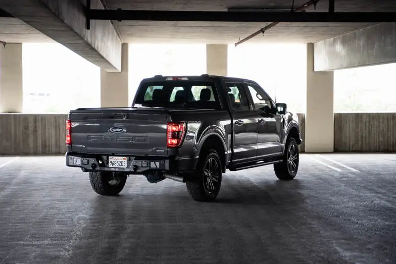 DV8 Offroad 21-23 Ford F-150 MTO Series Rear Bumper with steel construction and aluminum accents provides protection and utility steps, shown mounted on a gray Ford F-150 in a parking garage.