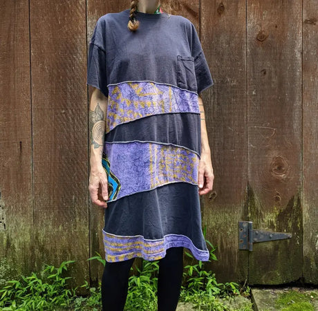 Person wearing a loose-fitting dress with horizontal purple and black strips, standing in front of a weathered wooden fence.