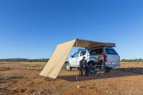 People sitting under ARB Wind Break-Side Fire Retardant awning attached to a parked silver pickup truck in a desert landscape.