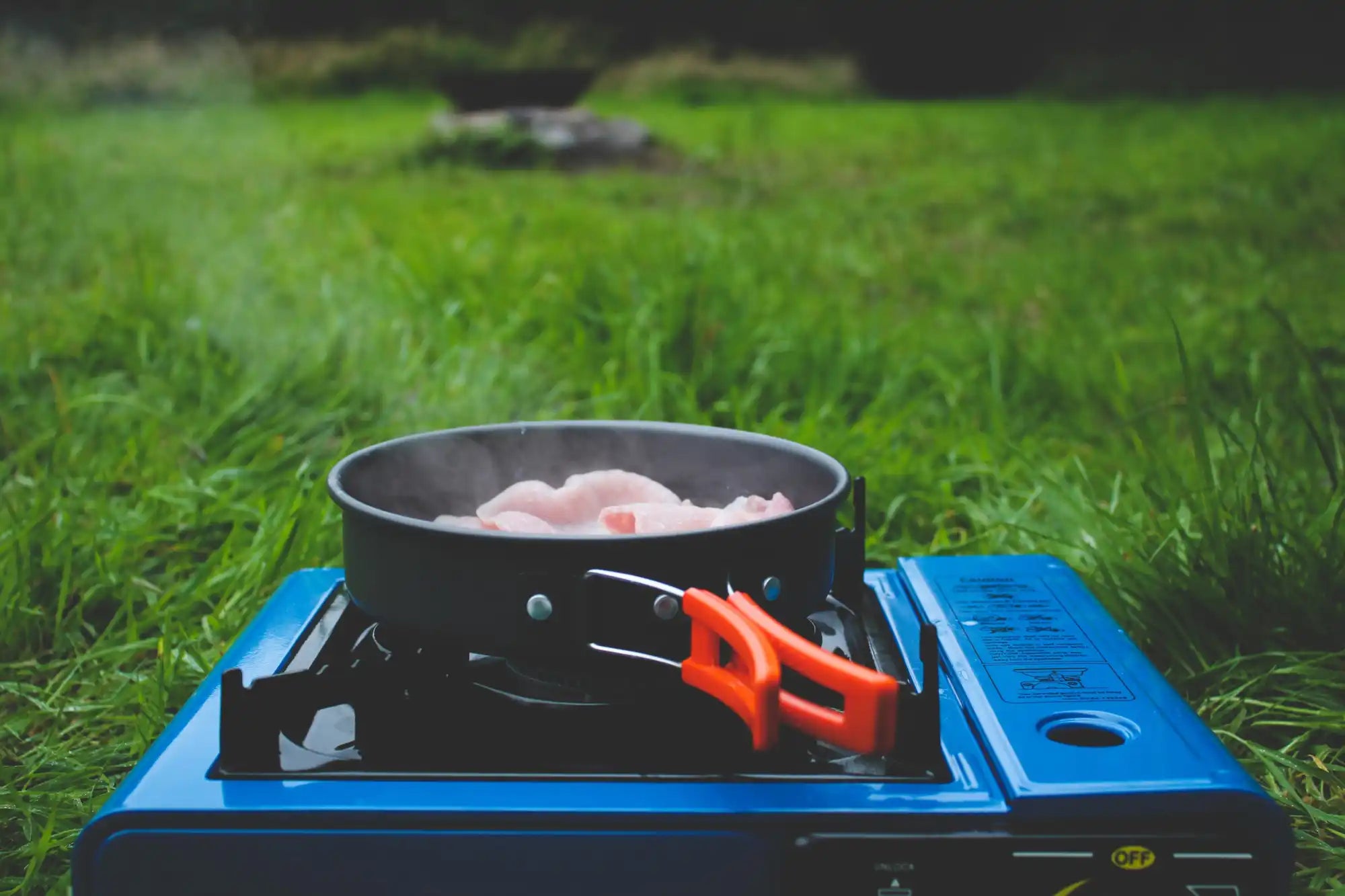 Blue camping stove with orange handle and black pot containing raw chicken.