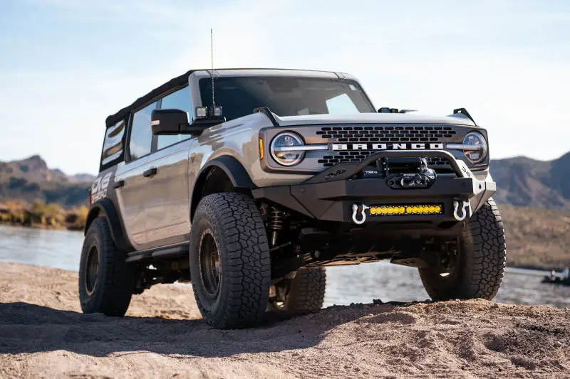 DV8 Offroad 2021+ Ford Bronco with Modular Front Bumper, Winch, and Auxiliary Light Mounts on rocky terrain with mountains in the background.