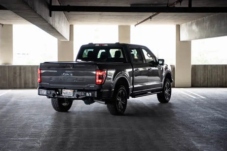 DV8 Offroad 21-23 Ford F-150 MTO Series Rear Bumper with steel construction and aluminum accents provides protection and utility steps, shown mounted on a gray Ford F-150 in a parking garage.