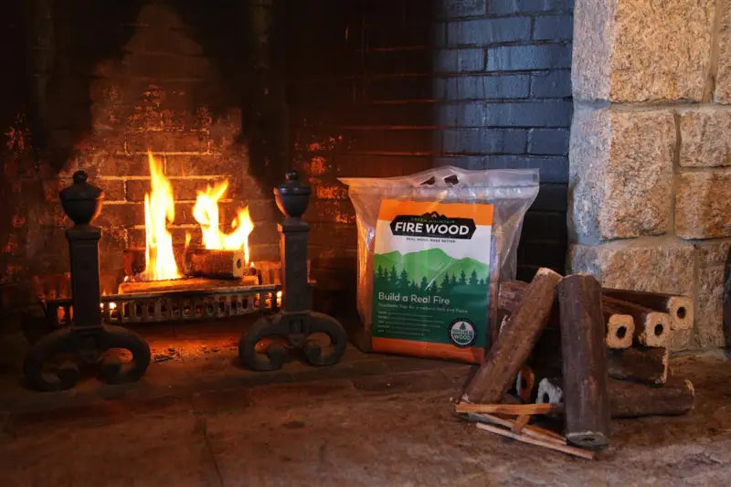 Fireplace with flames and burning Green Mountain Firewood from GMFW 8 PACK, a bag of firewood leaning against the back wall and additional logs piled next to it.